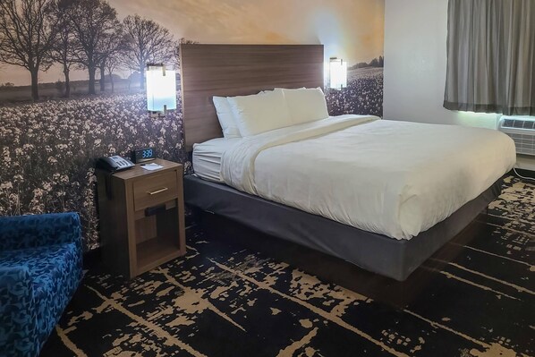 A hotel room with a large bed, a bedside table with a phone, a blue chair, and a patterned rug.