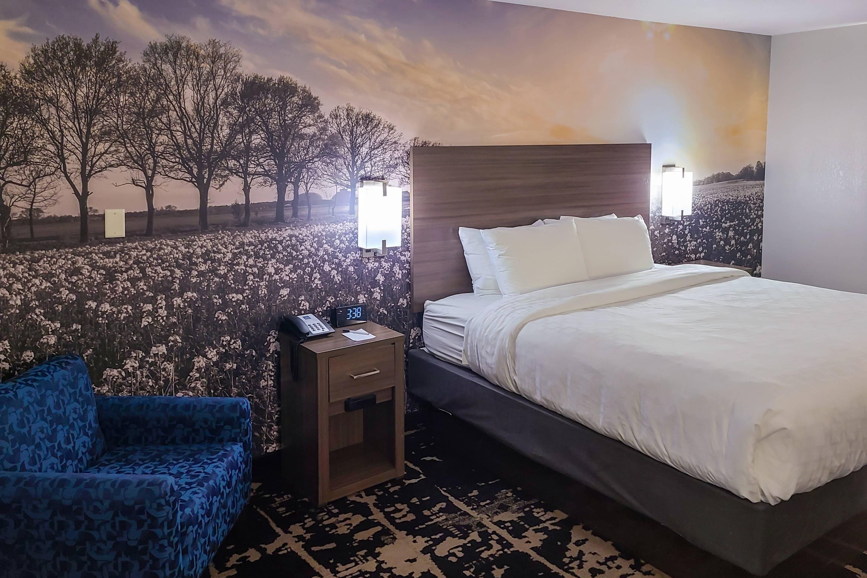 A hotel room with a bed, a nightstand with a phone, a blue chair, and a wall mural of trees and flowers.