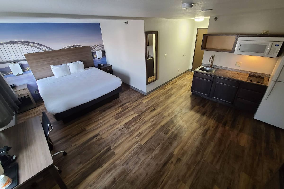 A hotel room with a bed, a desk, a mirror, a small kitchenette, and a view of a bridge.