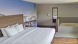 A hotel room with a large bed, a wooden floor, a bathroom with a bathtub, and a cityscape mural on the wall. - Clarion Pointe Bettendorf - Quad Cities (Bettendorf)