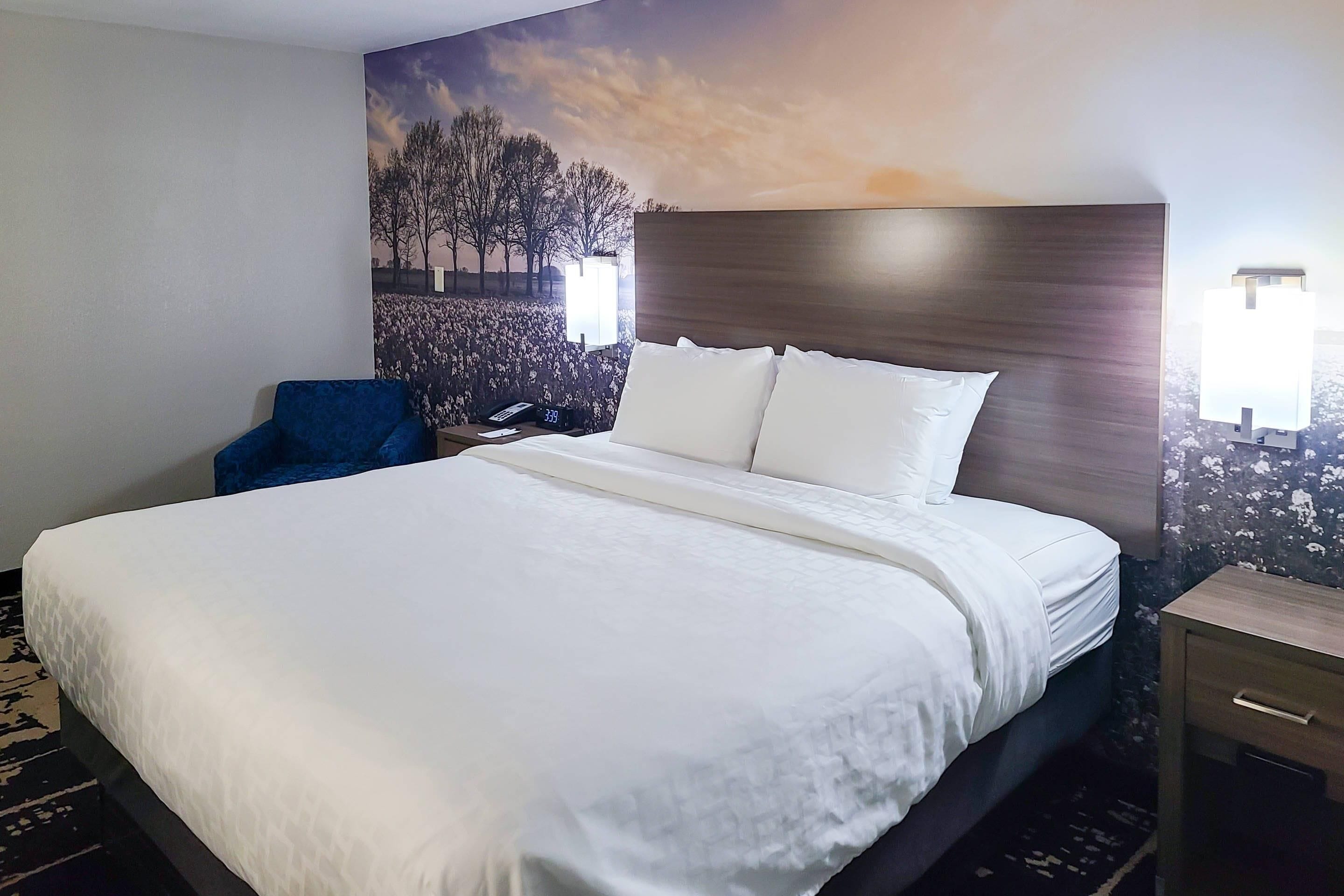 Habitación de hotel con una cama grande, una silla azul, una mesita de noche con una lámpara y un mural en la pared.