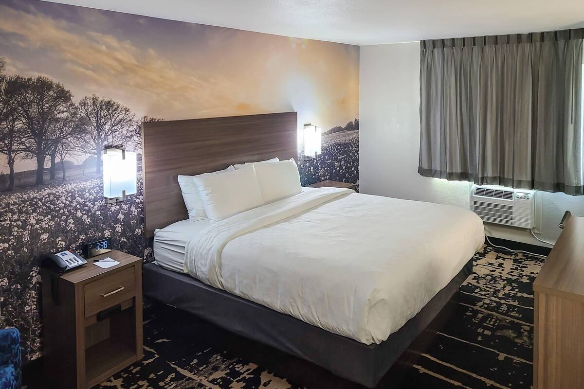 A hotel room with a large bed, a nightstand with a phone, a wall-mounted lamp, and a window with curtains.