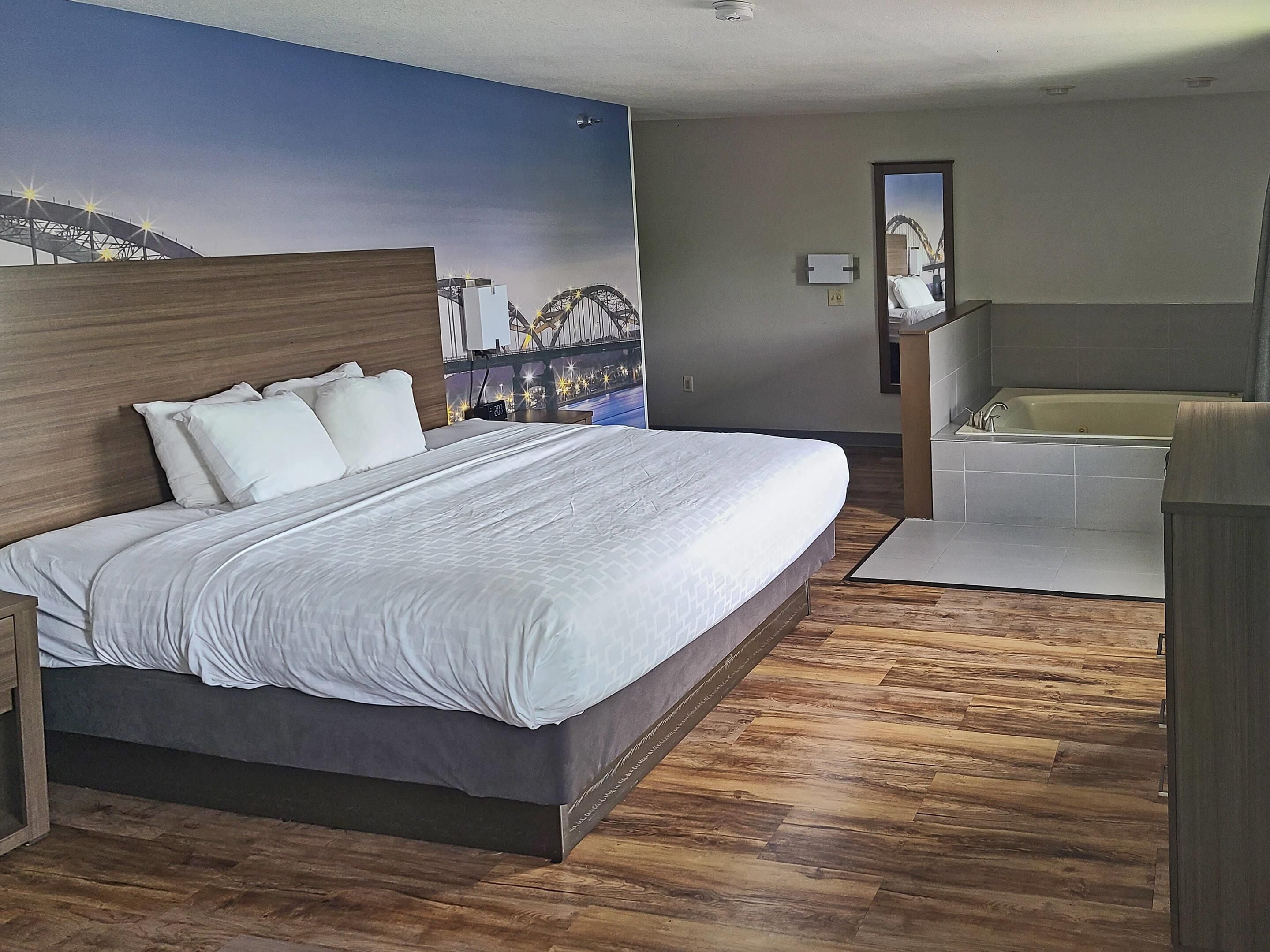 Ein Hotelzimmer mit einem großen Bett, einem Holz-Kopfstück, Wandbild einer Brücke, einem Badezimmer mit Badewanne und Holzboden.