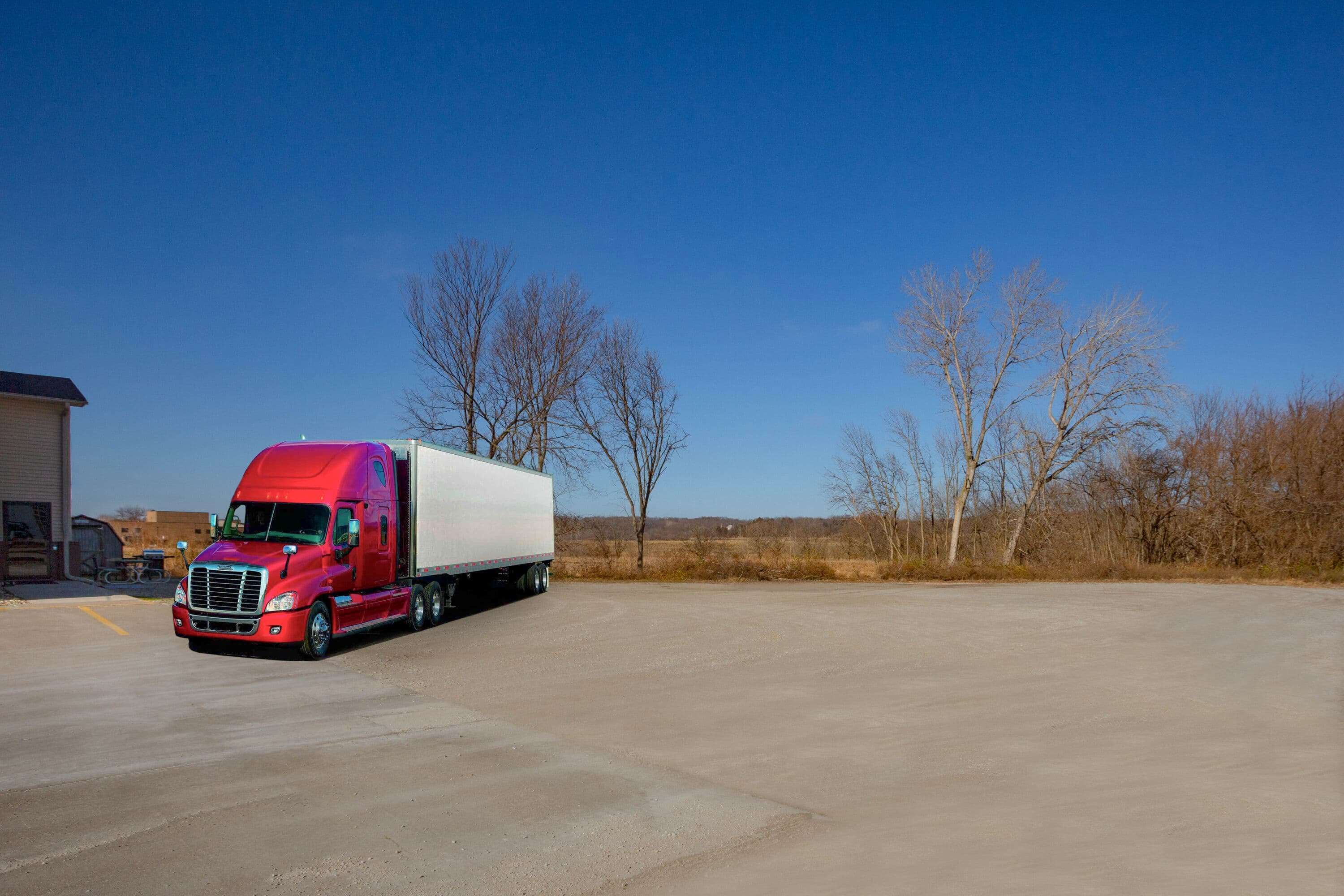 A red semi-truck parked in a lot with a clear blue sky and leafless trees in the background.