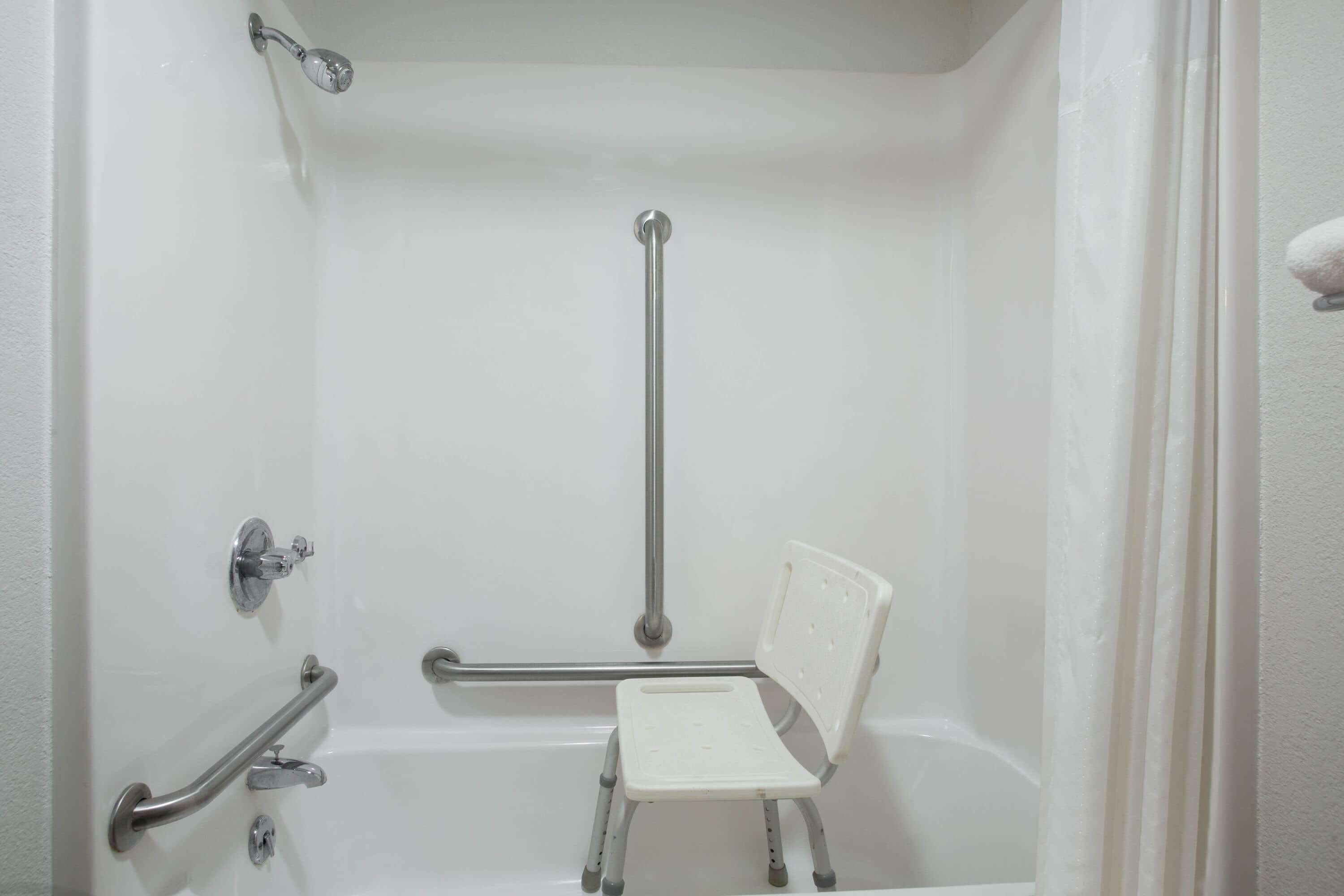 A bathroom with a white bathtub, grab bars, a shower chair, and a handheld showerhead.