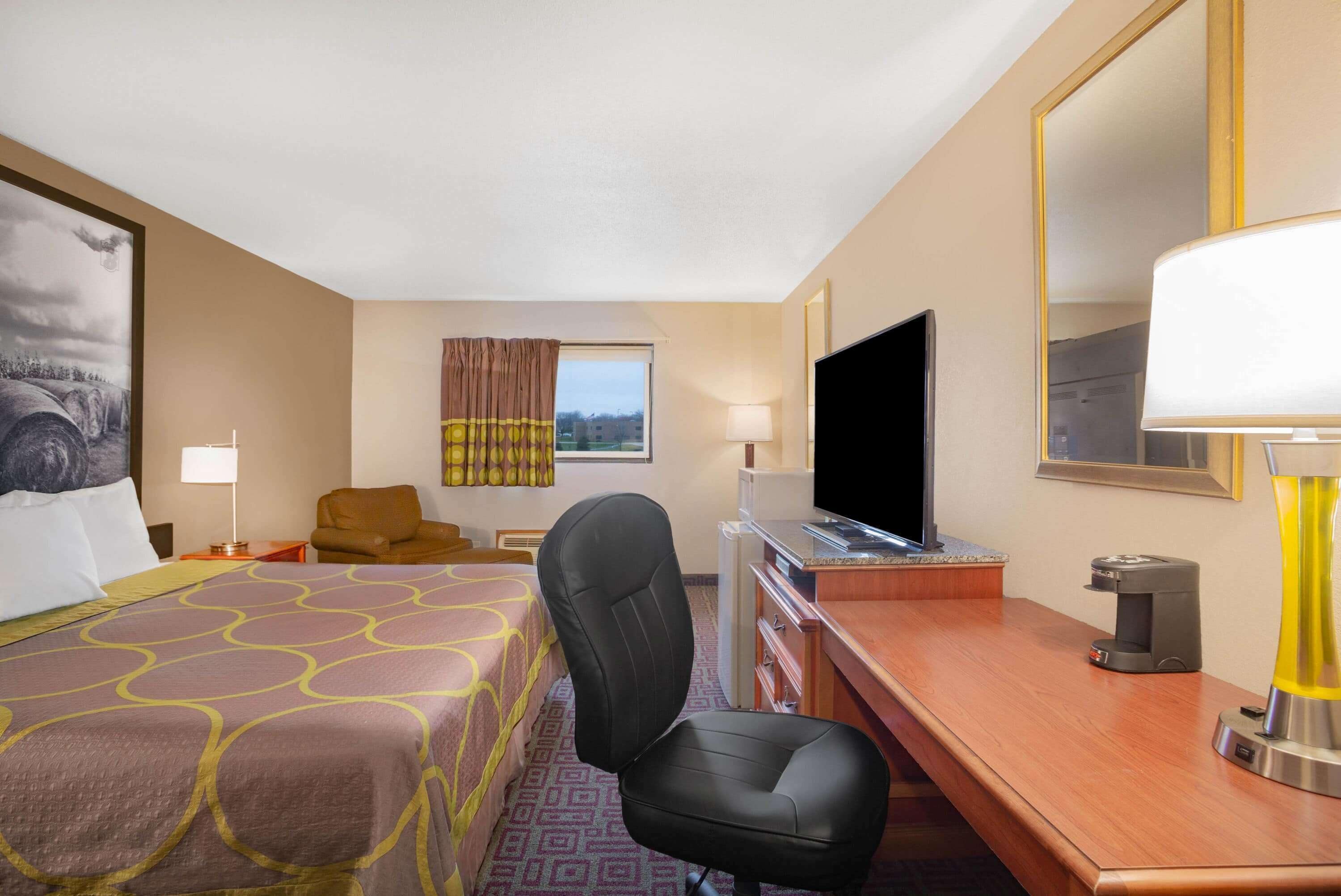 Quarto de hotel com cama, escrivaninha, cadeira, televisão, abajur e janela com cortinas.