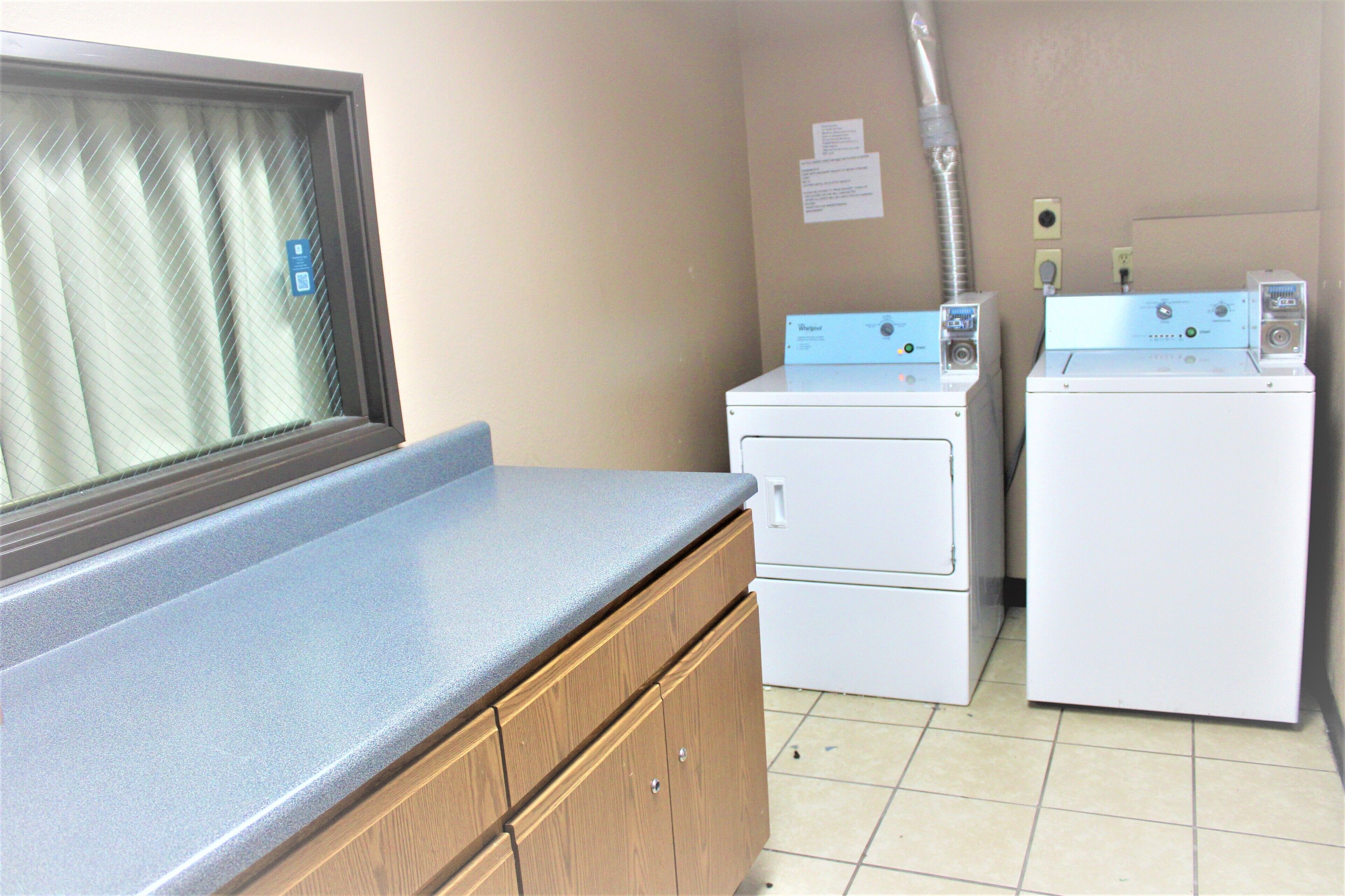 A laundry room with two washing machines, a countertop, and a window with blinds.