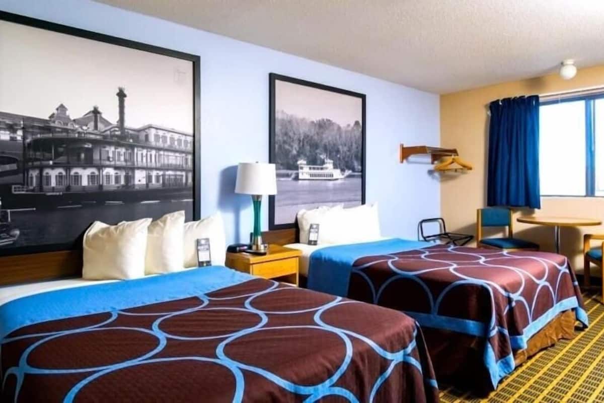 Ein Hotelzimmer mit zwei Betten, einem Schreibtisch, einem Sessel, einer Lampe und zwei gerahmten Bildern an der Wand.