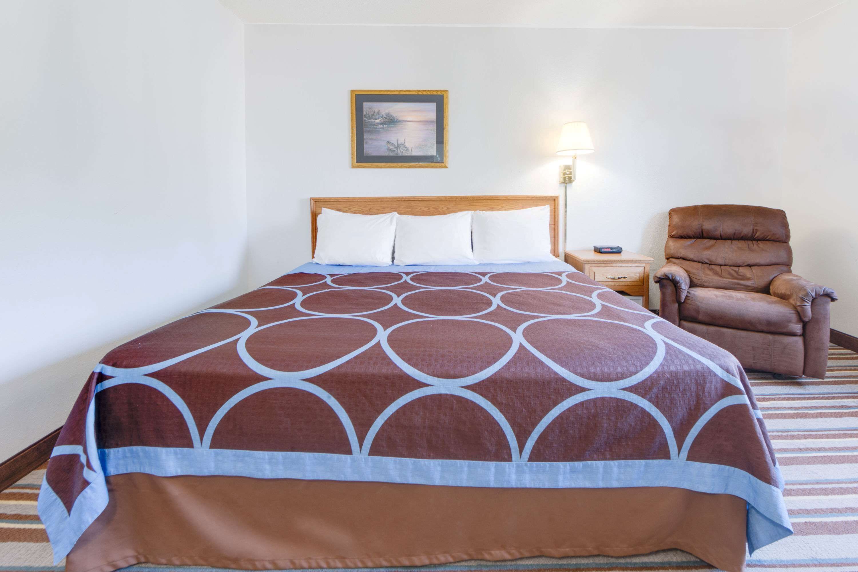 Een hotelkamer met een bed, een stoel, een nachtkastje, een lamp en een lijst met een foto aan de muur.