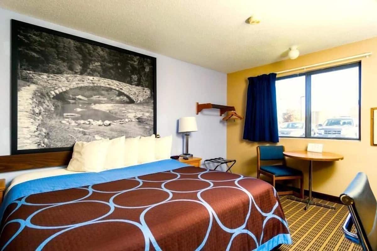 Ein Hotelzimmer mit Bett, Schreibtisch, Stuhl, Lampe, einem Fenster mit Vorhängen und einem großen Wandbild.