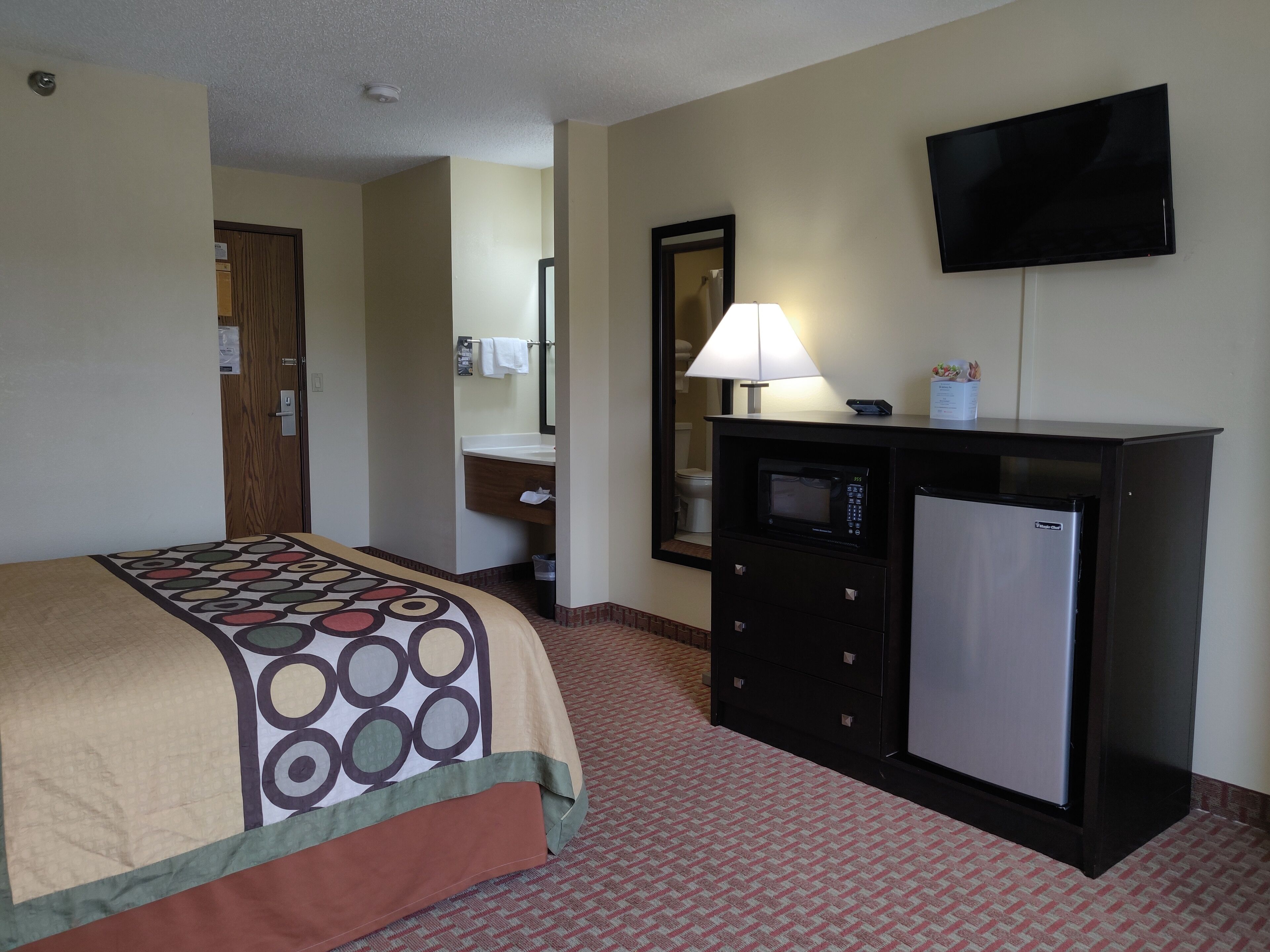 Habitación de hotel con cama, escritorio con microondas, refrigerador, televisor y baño a la vista a través de una puerta abierta.