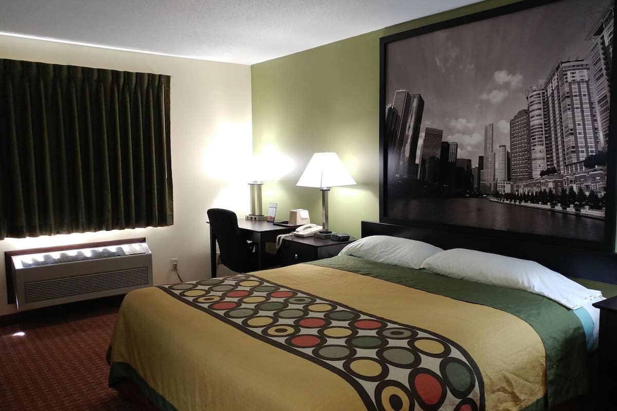 Hotelkamer met een bed, twee bedlampen, een bureau met telefoon, een stoel, een raam met gordijnen en een stadsgezicht schilderij aan de muur.