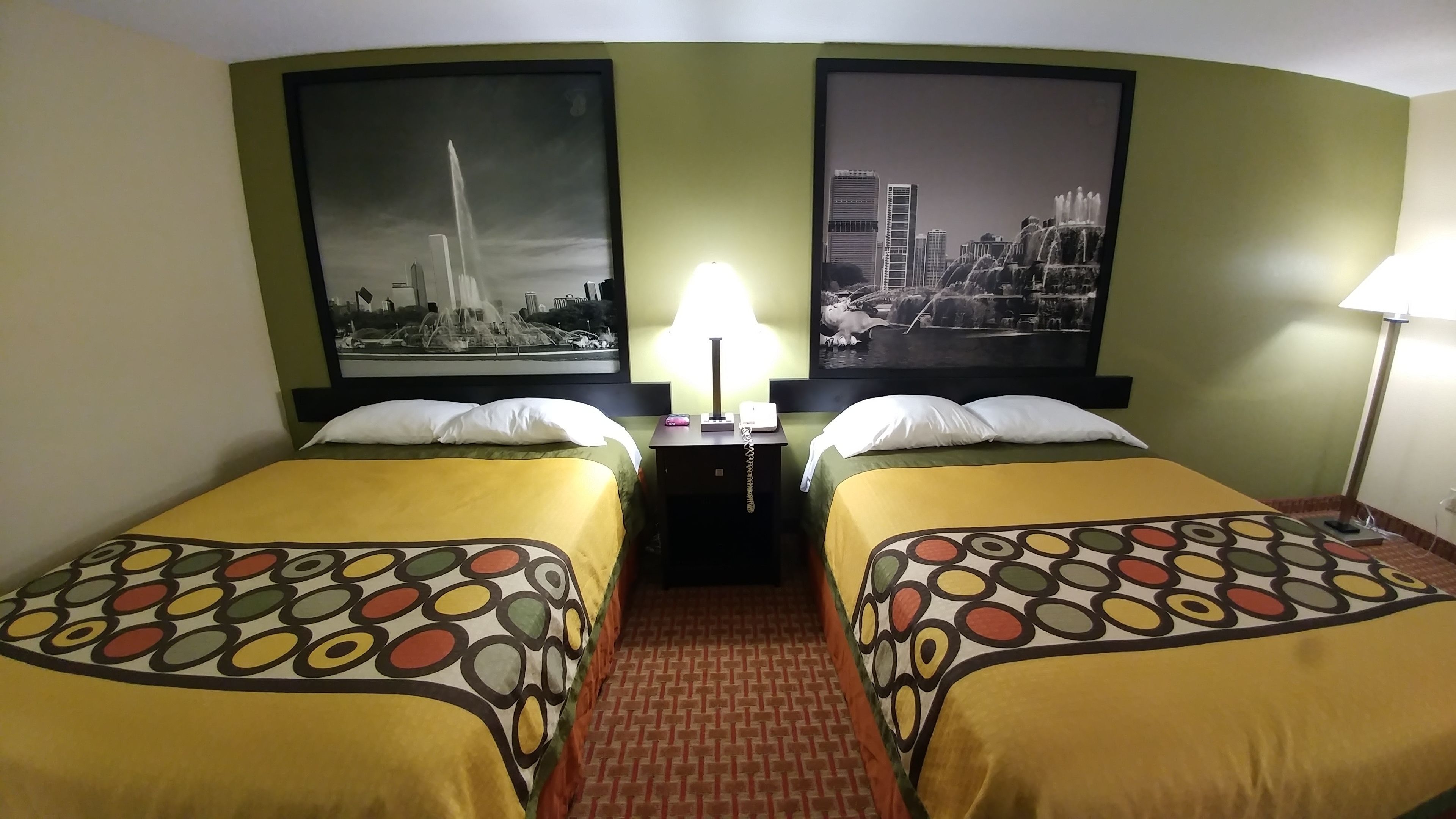 Habitación de hotel con dos camas, una mesita de noche, una lámpara y dos grandes carteles con vistas urbanas en las paredes.