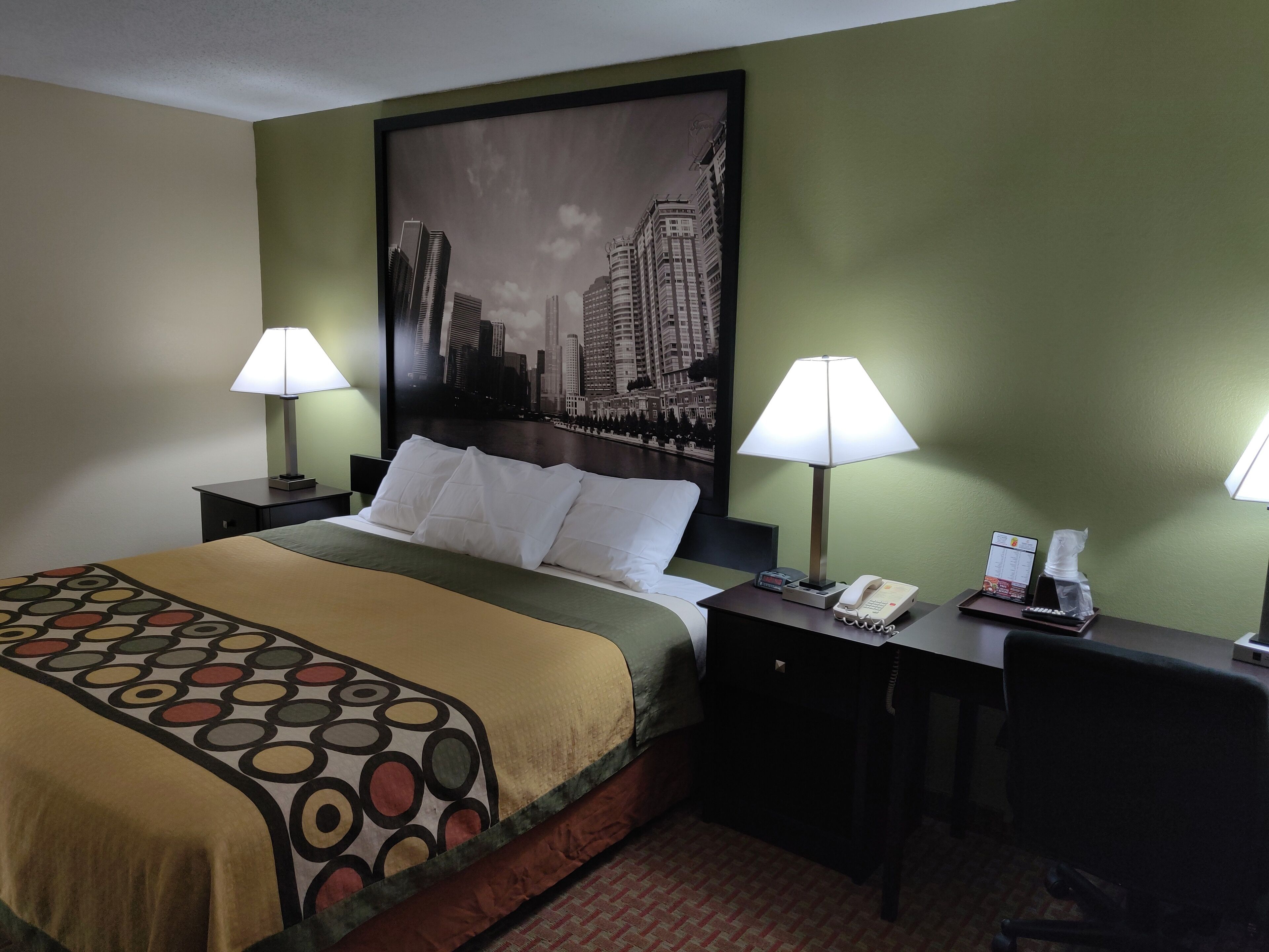 A hotel room with a bed, two bedside lamps, a nightstand with a phone and a menu, and a cityscape painting on the wall.