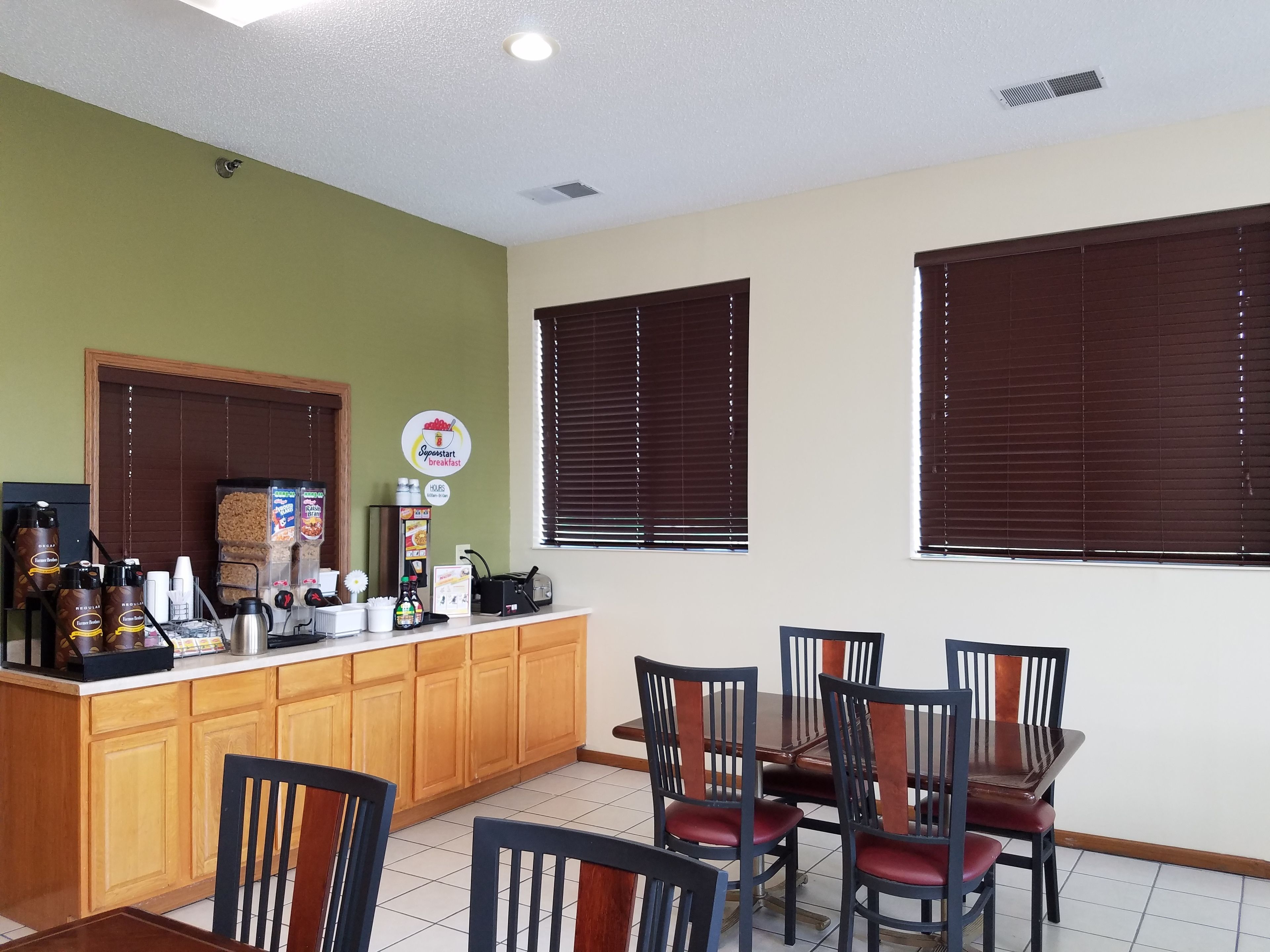 Una barra de desayuno con armarios de madera, una máquina de café y diversas opciones para el desayuno. Cuatro sillas rodean una mesa, y hay dos ventanas con persianas.