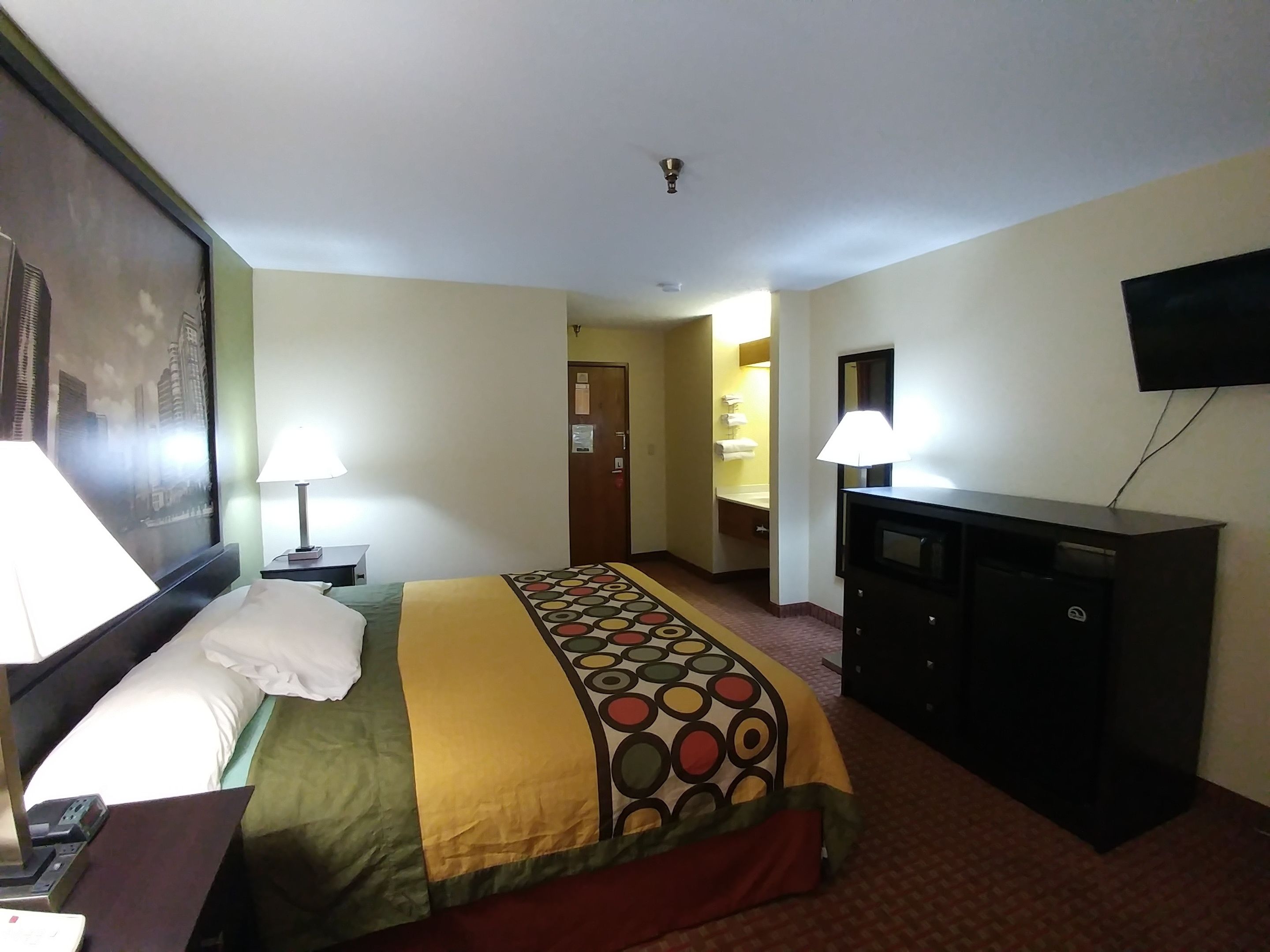A hotel room with a bed, two bedside lamps, a desk with a computer, a TV on a stand, and a bathroom visible through an open door.