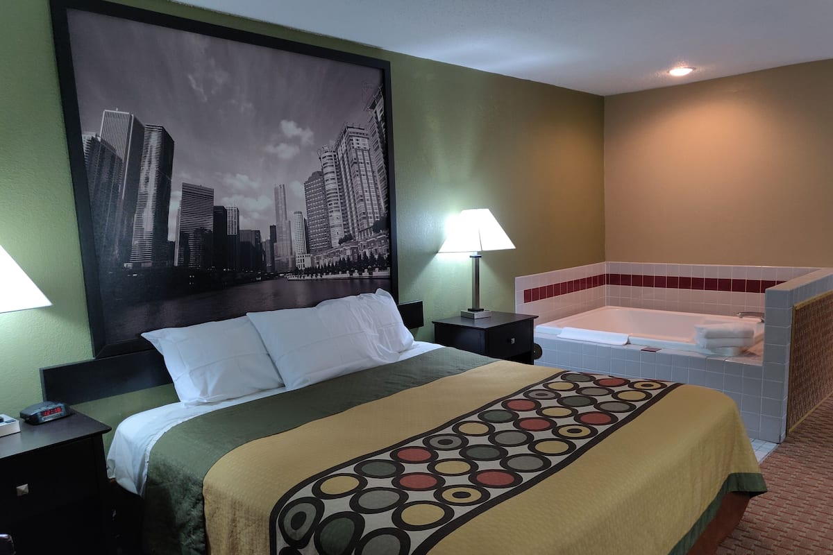 Hotelkamer met een bed, nachtkastje, lamp, groot stadslandschap schilderij en een bad.