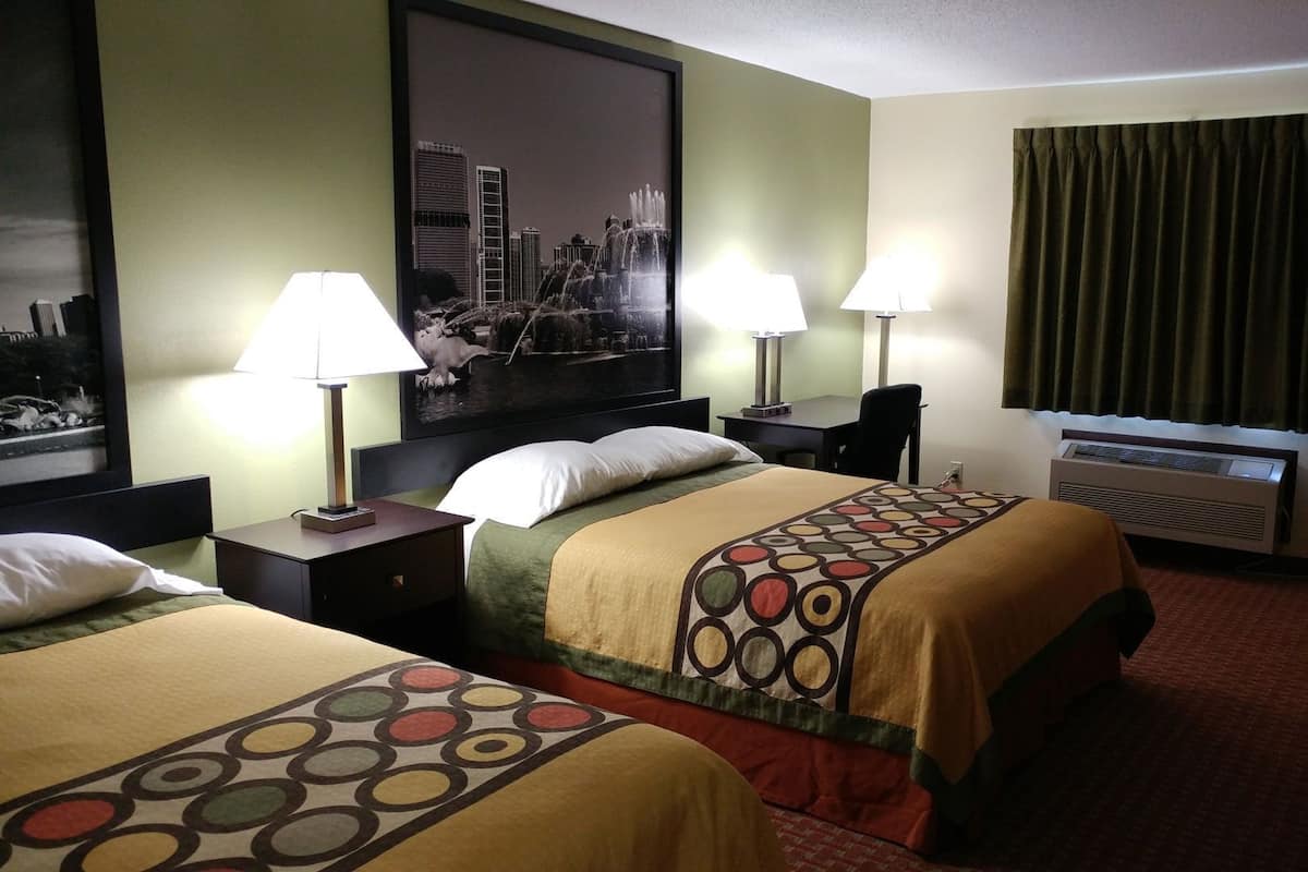 Een hotelkamer met twee bedden, een bureau, een stoel, een groot raam met gordijnen en een stadsgezichtschilderij aan de muur.