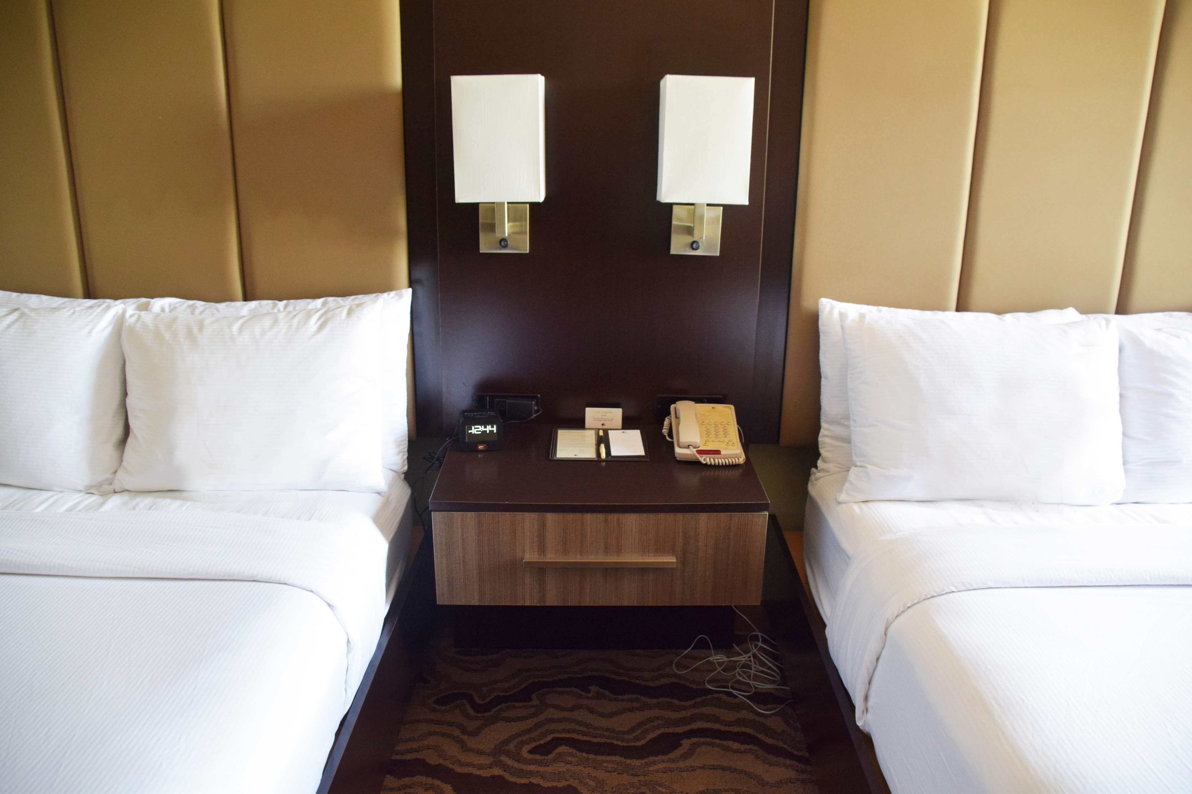 A hotel room with two beds, a nightstand with a lamp, a clock, a telephone, and a small drawer.