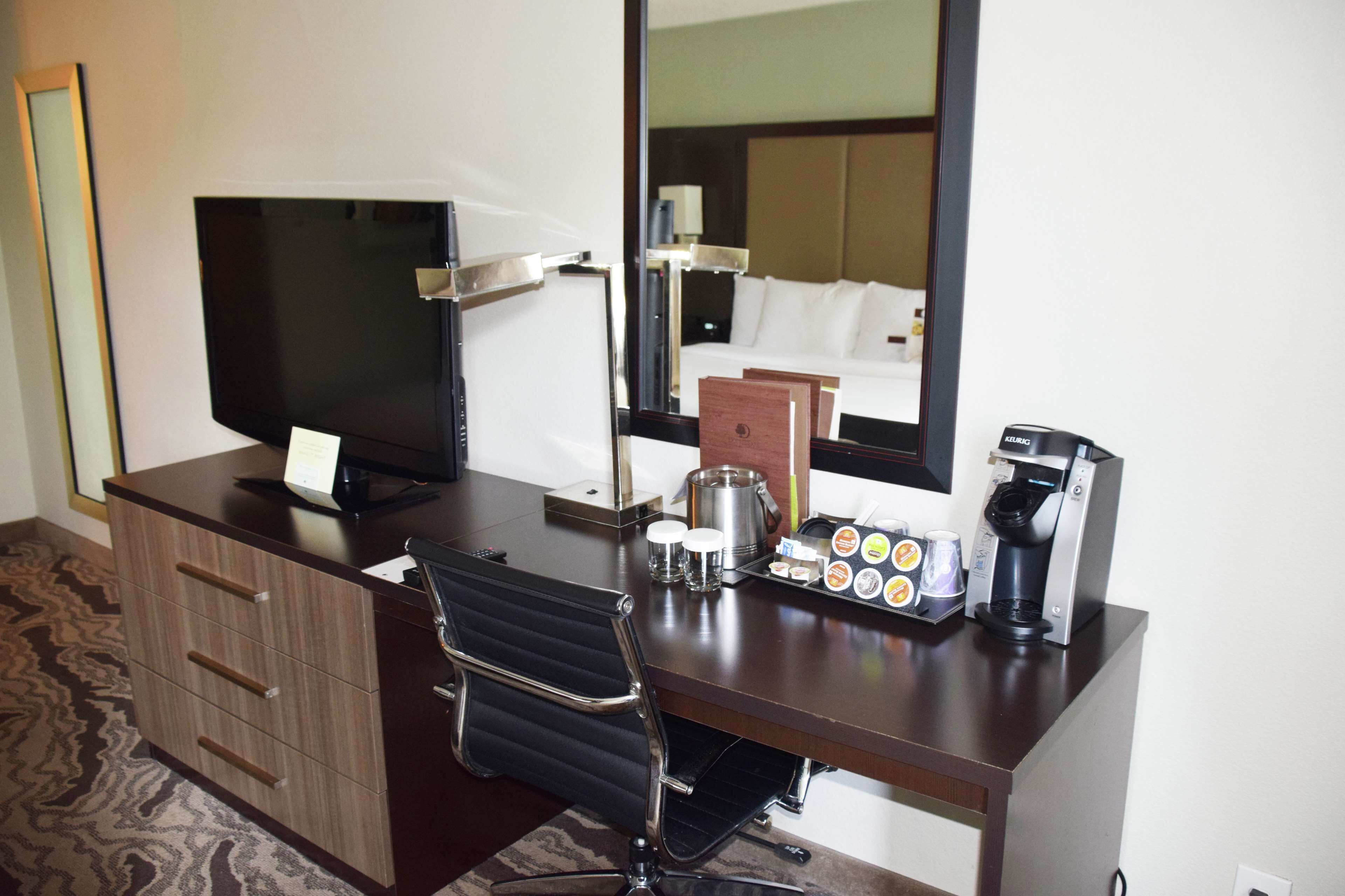 Une chambre d’hôtel équipée d’un bureau, d’une télévision à écran plat, d’une machine à café, d’un miroir et d’un lit avec du linge de lit blanc.