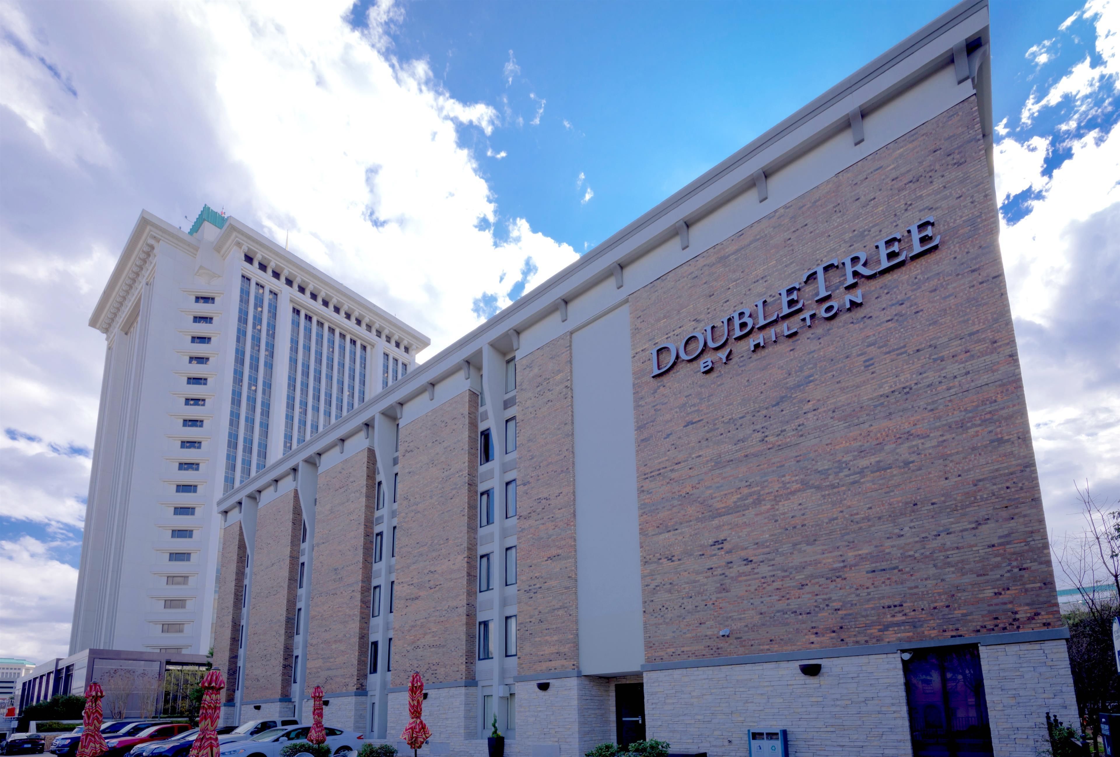 A hotel with a prominent "DOUBLE TREE by Hilton" sign, a tall building with multiple windows, and a parking area with cars and umbrellas.