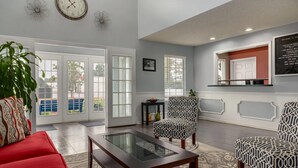 A spacious living area with a red sofa, patterned armchairs, a glass coffee table, and a large window offering a view of the outdoors. - Baymont by Wyndham Albany at Albany Mall (Albany)