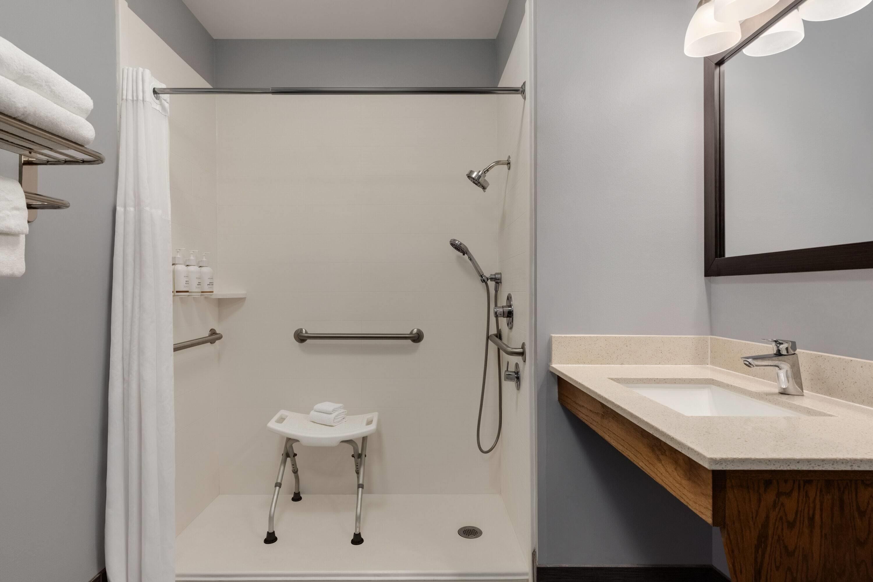 A modern bathroom with a walk-in shower, grab bars, a small sink, and a mirror.