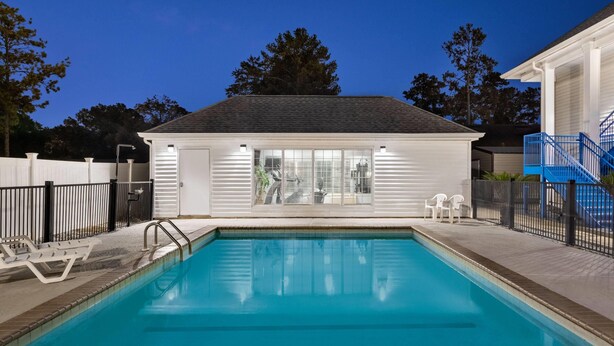 A rectangular swimming pool with a tiled deck, surrounded by a white fence, and a small building with large windows in the background. - Baymont by Wyndham Albany at Albany Mall (Albany)
