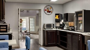 A modern kitchen with dark wood cabinets, a microwave, coffee maker, and a refrigerator. There is a dining area with blue chairs and a table. - Baymont by Wyndham Albany at Albany Mall (Albany)