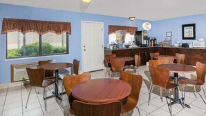 A restaurant with round tables and chairs, a kitchen area, and a view of greenery through the windows.