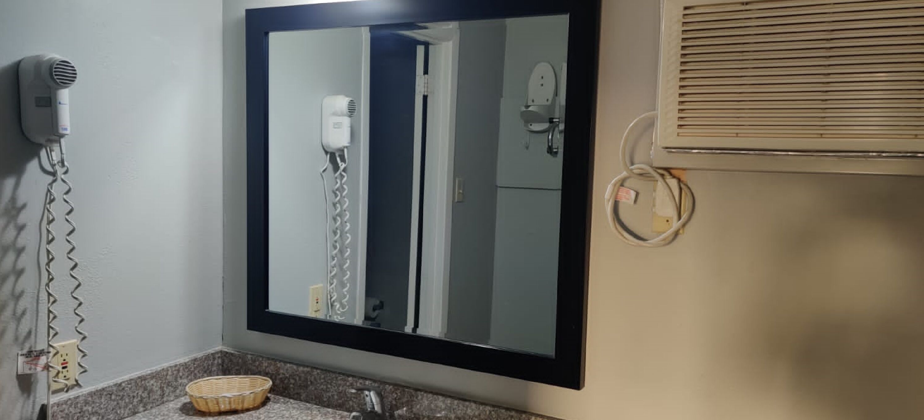 A bathroom with a hairdryer mounted on the wall, a mirror, and a window with blinds.