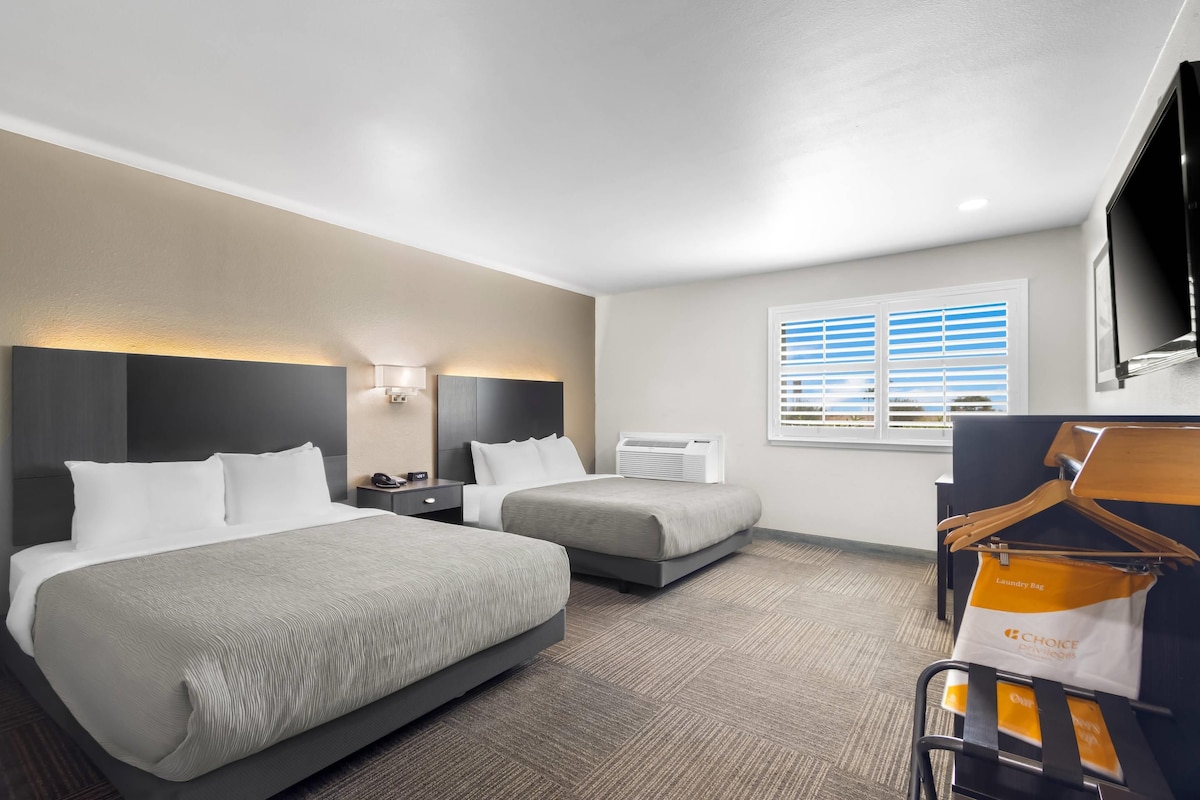 A hotel room with two beds, a flat-screen TV, a window with blinds, and a luggage cart with a 'Choice Privileges' bag.