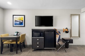 A hotel room with a flat-screen TV mounted on the wall, a minibar, a desk with a chair, and a mirror.