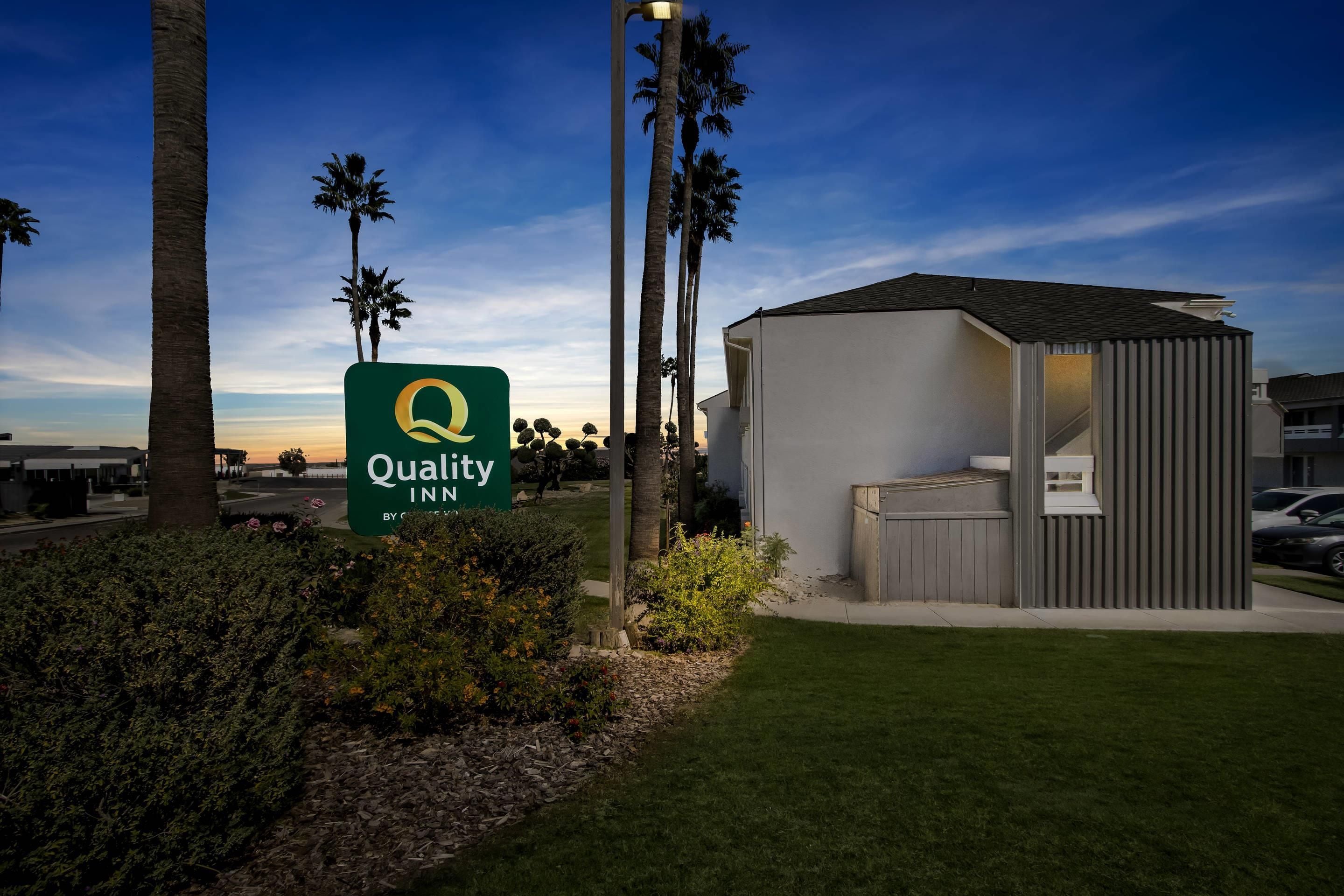 A Quality Inn sign, a small building, and palm trees.