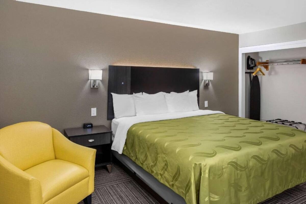 Ein Hotelzimmer mit einem Bett, einem Nachttisch, einem Sessel, einem Kleiderhaken und einer Wandleuchte.