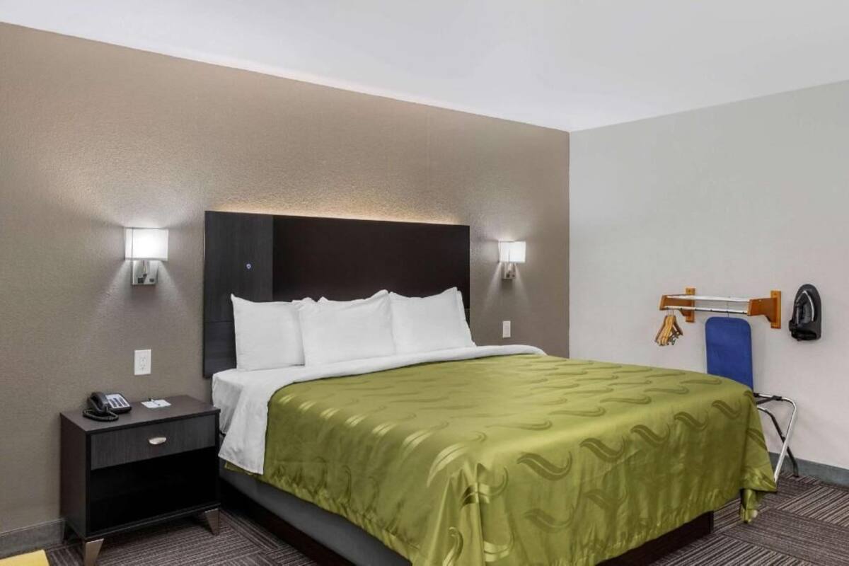 Ein Hotelzimmer mit einem Bett, Nachttischlampen, einem Telefon, einem Sessel und einem an der Wand montierten Bügelbrett.