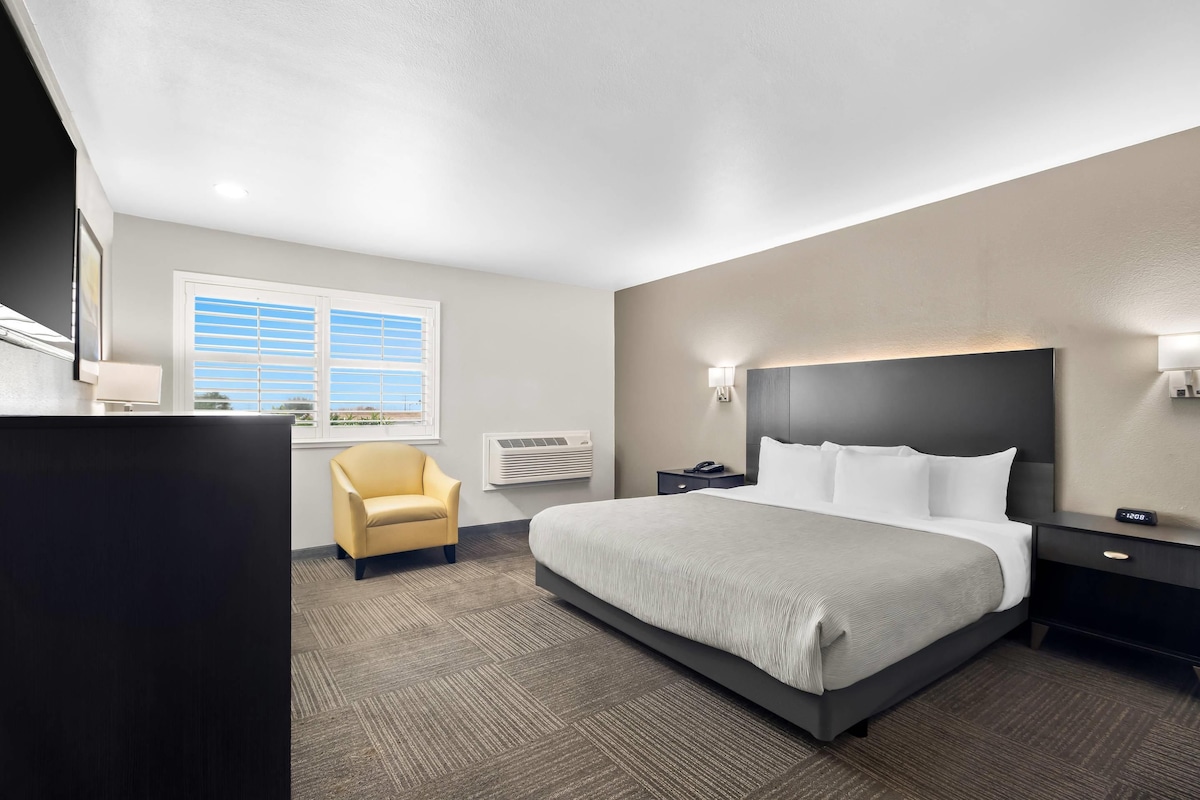 A hotel room with a large bed, a desk, a chair, a nightstand, a wall-mounted lamp, and a window with blinds.