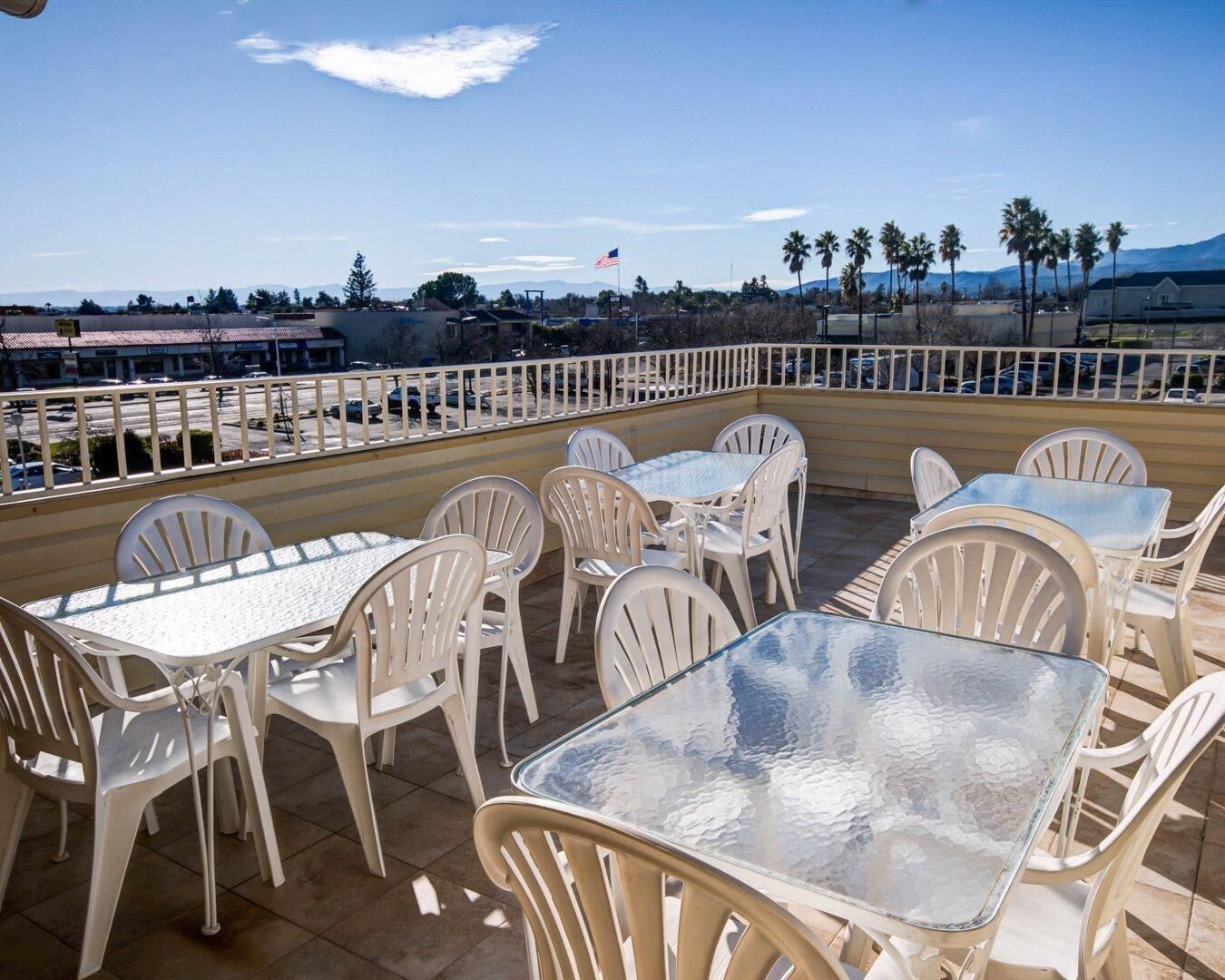 Eine Außenpatio mit weißen Plastikstühlen und -tischen, einem weißen Geländer und Blick auf einen Parkplatz sowie Berge im Hintergrund.