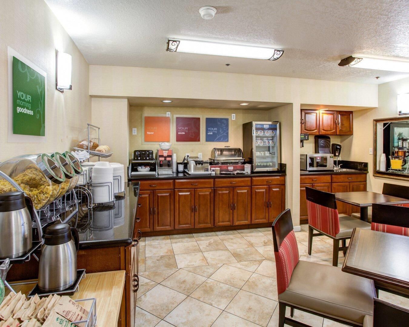 A breakfast bar with coffee machines, a refrigerator, and a microwave. There are wooden cabinets and a tiled floor.