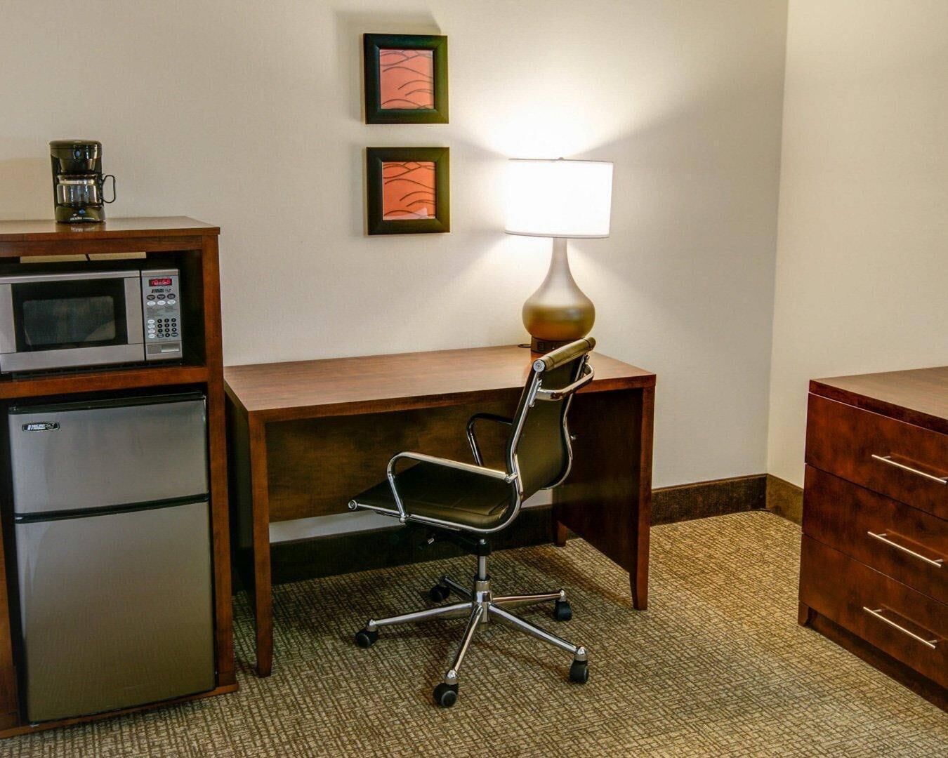Habitación de hotel con un escritorio de madera, una silla, una lámpara, un microondas y un refrigerador.