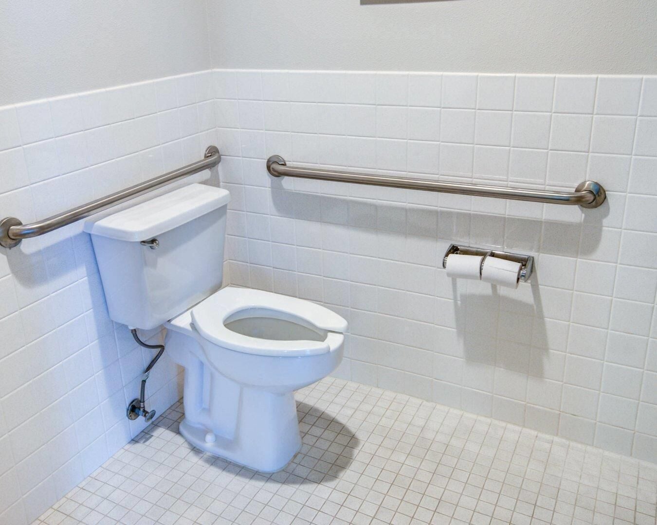 Une salle de bain avec des murs recouverts de carreaux blancs, des toilettes blanches et des barres d’appui installées pour plus de sécurité.