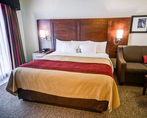 A hotel room with a large bed, a nightstand, a lamp, a chair, and a framed picture on the wall.