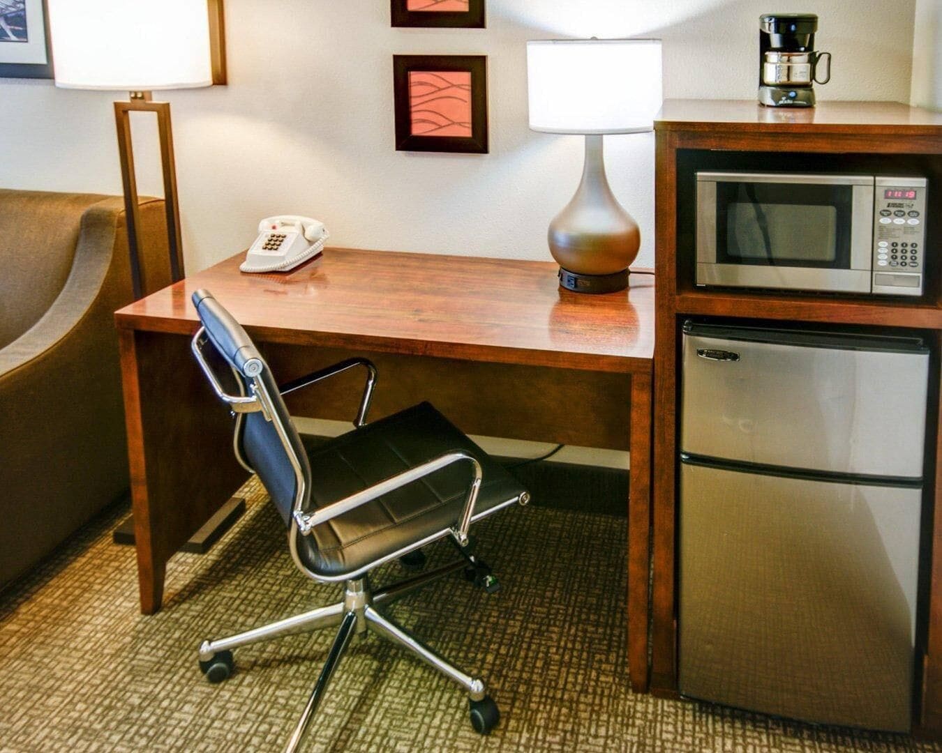 A hotel room with a wooden desk, a black office chair, a telephone, a lamp, a microwave, and a refrigerator.