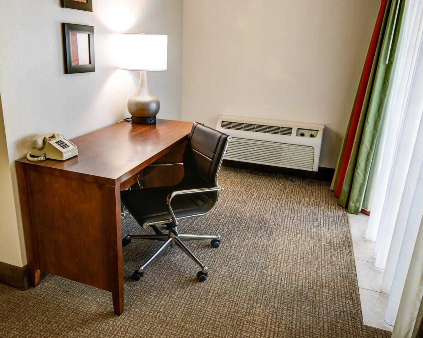 Habitación de hotel con escritorio de madera, silla de oficina negra, lámpara, teléfono y calefactor en la pared.