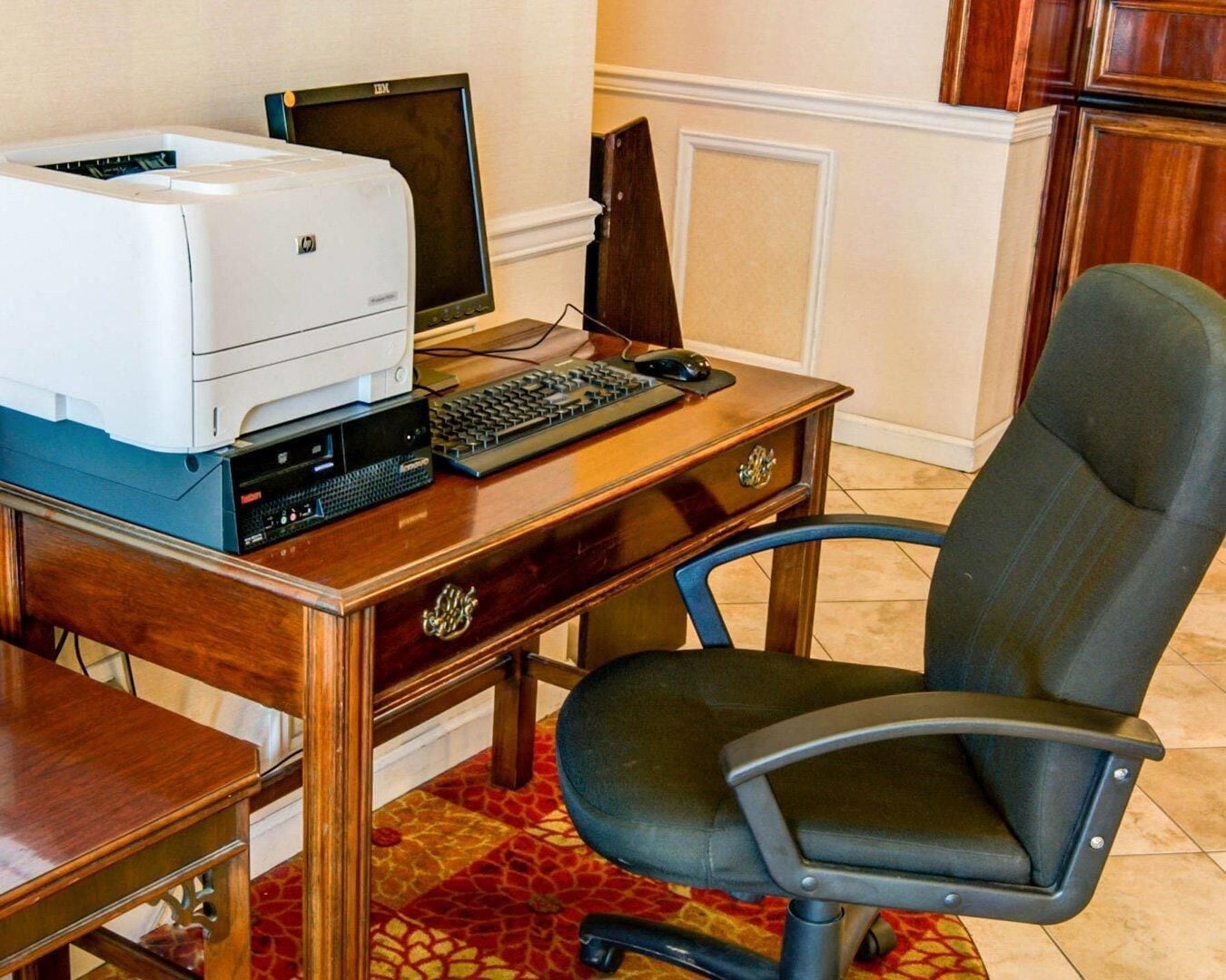 A wooden desk with a printer, a computer monitor, a keyboard, and a chair.