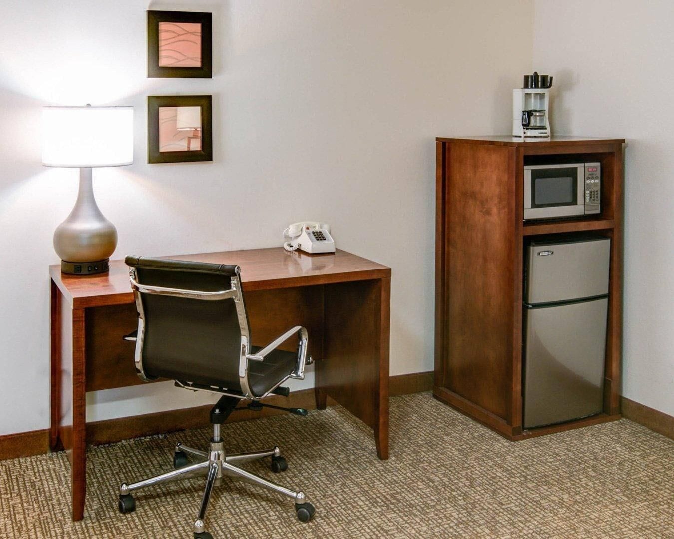 A hotel room with a wooden desk, a black office chair, a lamp, a telephone, a microwave, and a refrigerator.