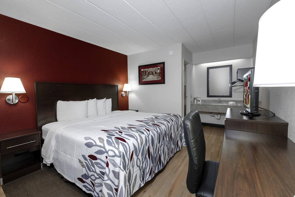 A hotel room with a large bed, a desk with a computer, a chair, a nightstand, a lamp, and a framed picture on the wall.
