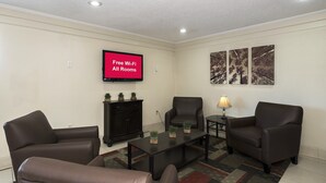 A furnished living room with a flat-screen TV displaying 'Free Wi-Fi All Rooms,' a dark wooden cabinet, two armchairs, a coffee table, and a lamp. - Red Roof Inn Bloomington - Normal/ University (Bloomington)