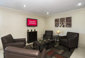 A furnished living room with a flat-screen TV displaying 'Free Wi-Fi All Rooms,' a dark wooden cabinet, two armchairs, a coffee table, and a lamp. - Red Roof Inn Bloomington - Normal/ University (Bloomington)