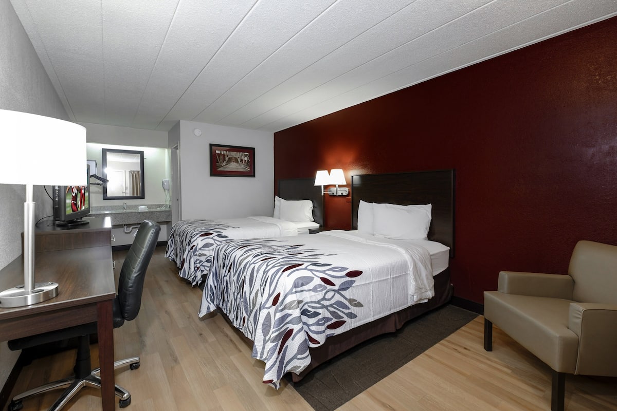A hotel room with two beds, a desk, a chair, and a desk lamp.